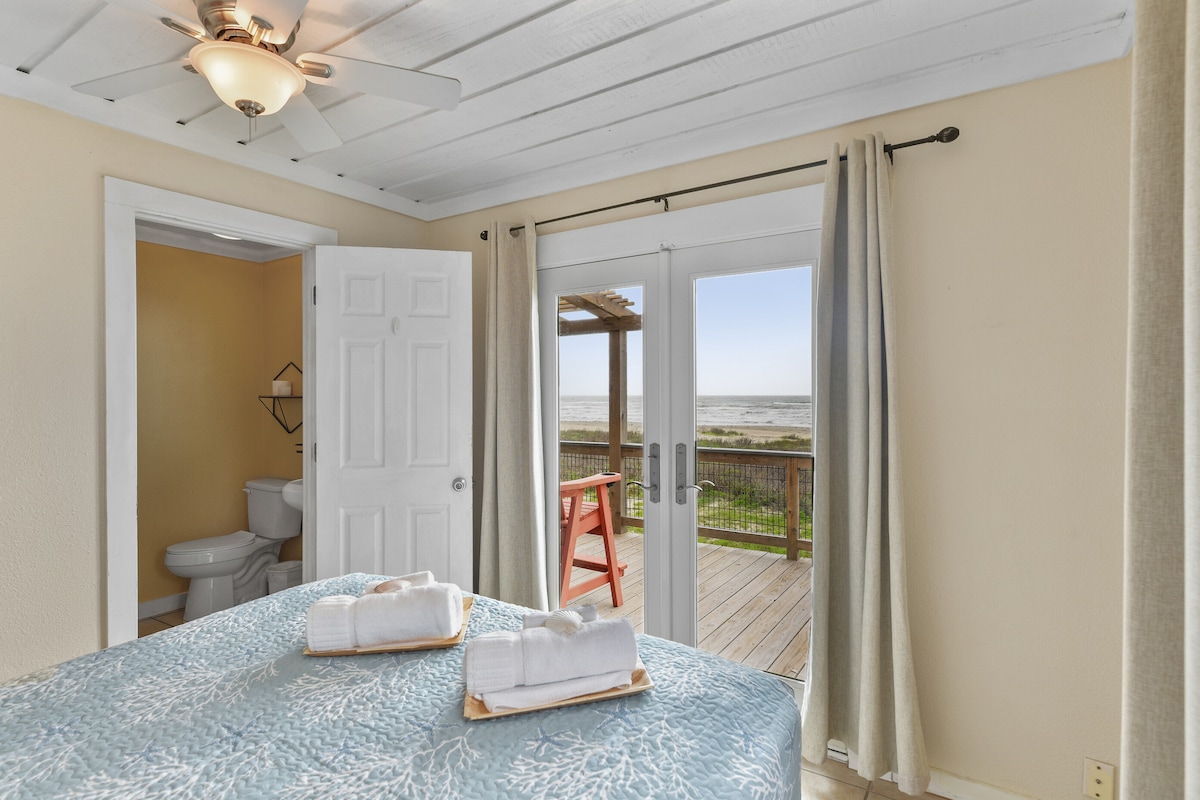 A bright bedroom showcases a spacious bed adorned with a light blue coverlet, paired with fluffy white towels. Double doors open to a private deck with views of the beach. A doorway leads to an ensuite bathroom, visible in the background.