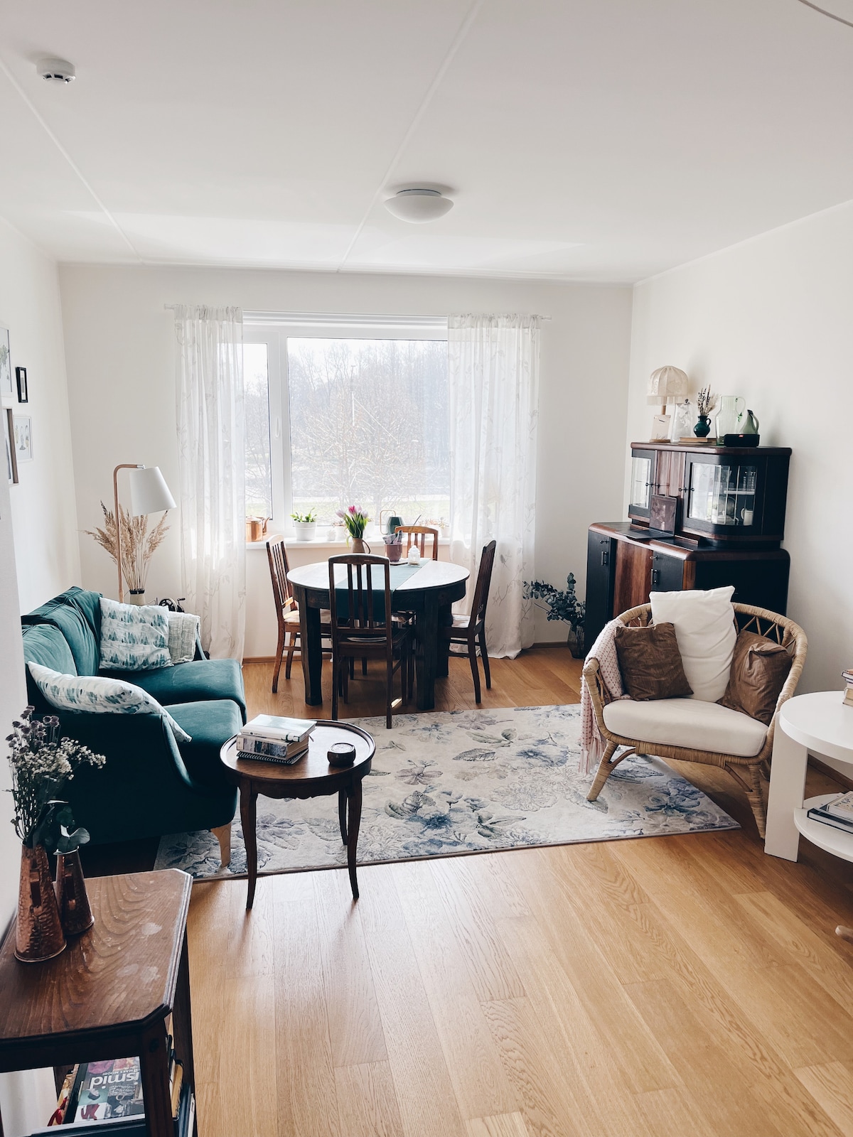 A welcoming living area showcases a combination of comfortable seating options including a green couch and a rocking chair. A round dining table is positioned next to a window, allowing light to fill the space. Decorative elements and a patterned rug enhance the cozy ambiance.