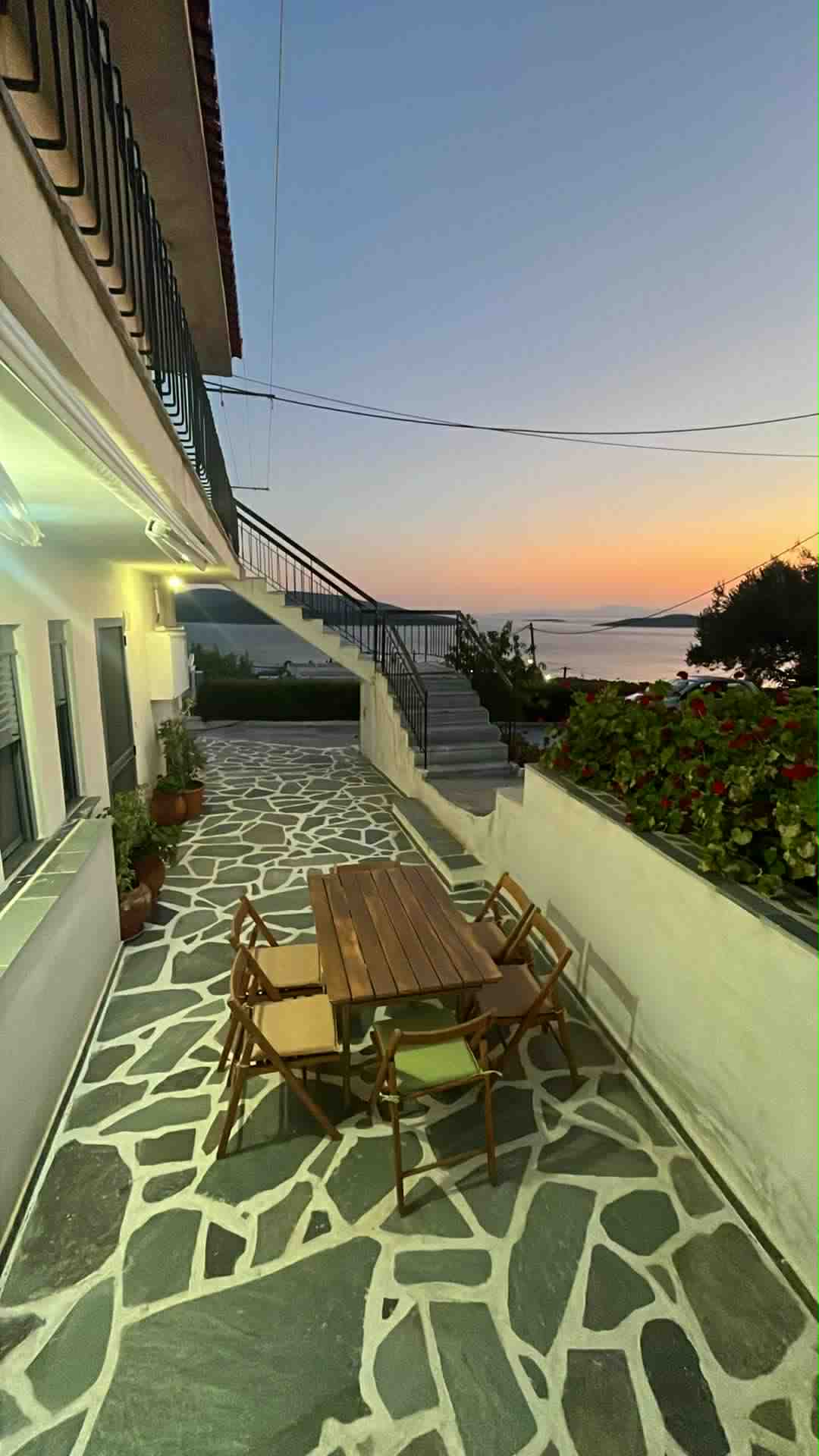 An outdoor dining area is shown, featuring a wooden table surrounded by several chairs. A stone pathway leads to a staircase, with potted plants lining the walls. A view of the serene sea and a colorful sky during dusk is visible in the background.