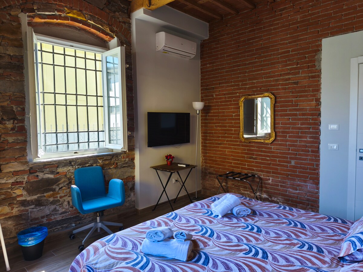 A cozy bedroom features a spacious bed adorned with a patterned bedspread. Natural light enters through a window, which is complemented by a decorative mirror and a flat-screen TV. A small table and a blue chair are present, along with exposed brick walls enhancing the room's character.