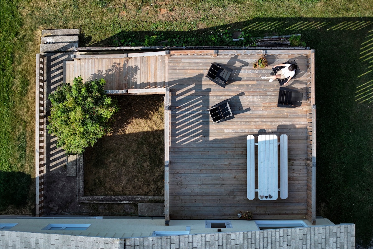 An elevated view of the outdoor patio features a wooden deck with several lounge chairs and a dining table. A small garden area is visible alongside a shaded tree, contributing to a relaxed outdoor space.