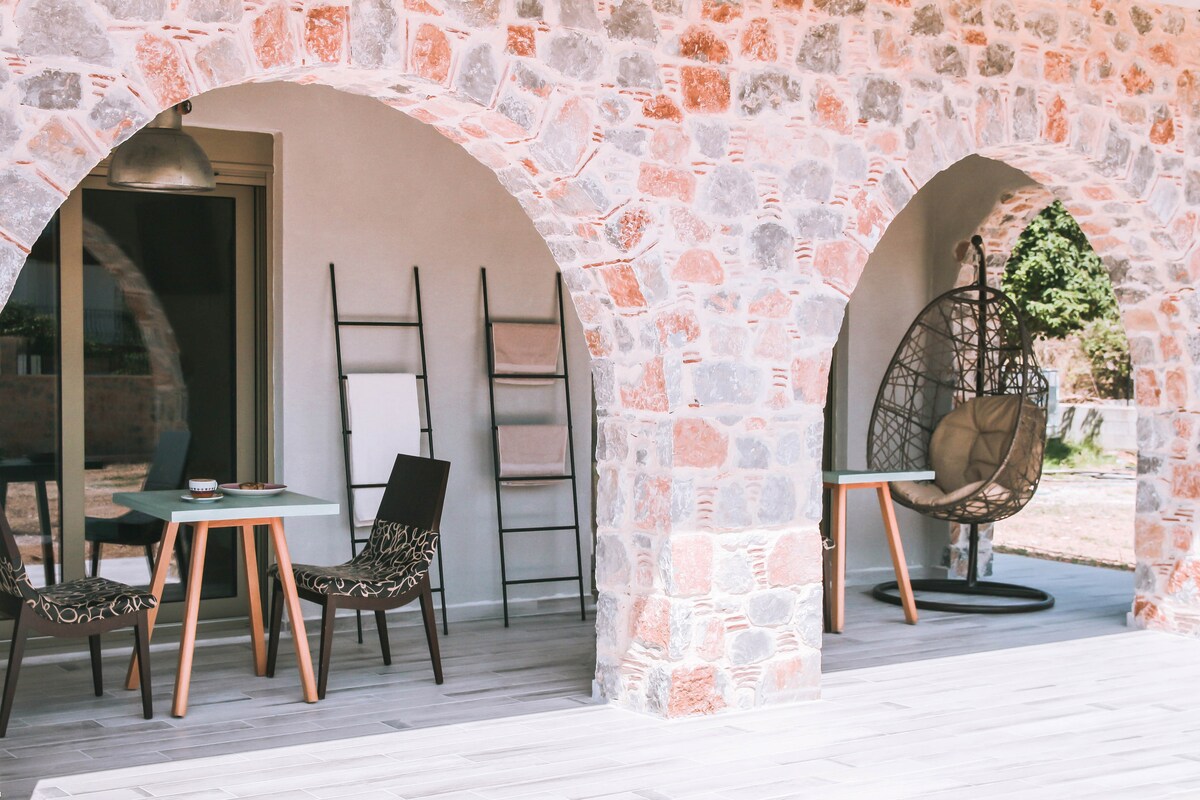 A charming outdoor area features two archways made of natural stone. One arched entrance leads to a dining table with chairs, while the other showcases a cozy hanging chair. A leaning ladder adds an element of design, complementing the relaxed atmosphere.
