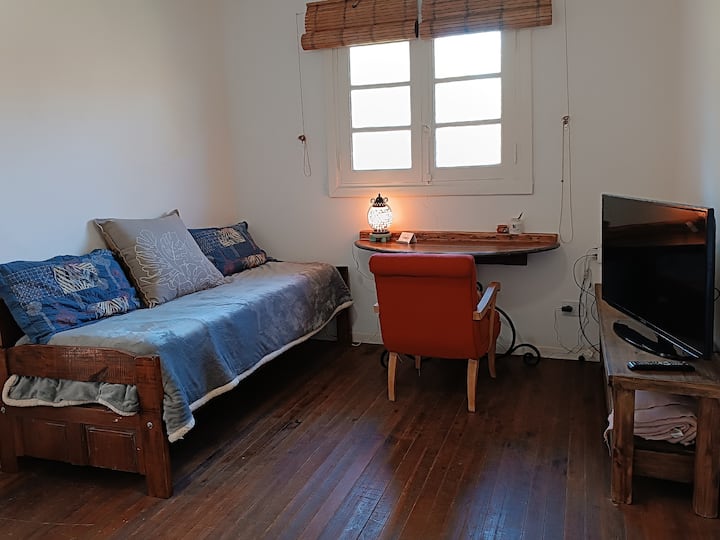 غرفة معيشة داخلية بسرير واحد ومكتب وتلفزيون ذكي مع جهاز بث (جهاز كروم كاست 4) وواي فاي وتدفئة بواسطة بلافا ومروحة سقف وإطلالة على الحديقة.