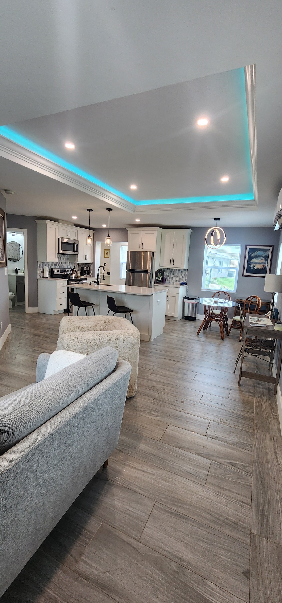An open living area is highlighted by modern furnishings and a light color palette. A spacious kitchen features stainless steel appliances and a bar with seating. Natural light fills the room through large windows, while recessed lighting adds a soft glow to the ceiling.