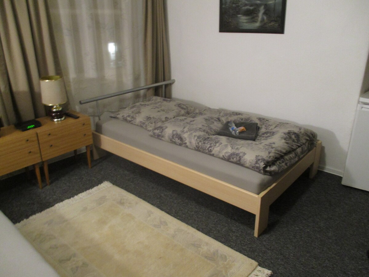 A cozy room features a single bed with a grey mattress and patterned bedding. A nightstand with a lamp and clock is positioned beside the bed. A small fridge is visible in the corner, while soft curtains frame the window, allowing gentle light to enter.