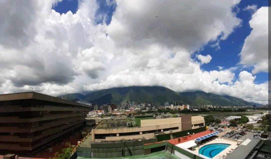 A panoramic view showcases a vibrant cityscape with mountains in the background. Generous natural light highlights the modern buildings and a clear blue sky dotted with fluffy clouds. The image captures both urban and natural elements, creating a serene atmosphere.