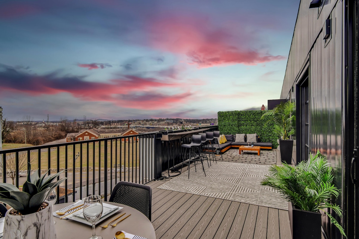 A spacious terrace is shown with a modern design, featuring an outdoor dining area and comfortable seating. Lush greenery and potted plants complement the setting, while the skyline is highlighted against a colorful sunset sky. The expansive view of Nashville's landscape can be seen in the background.