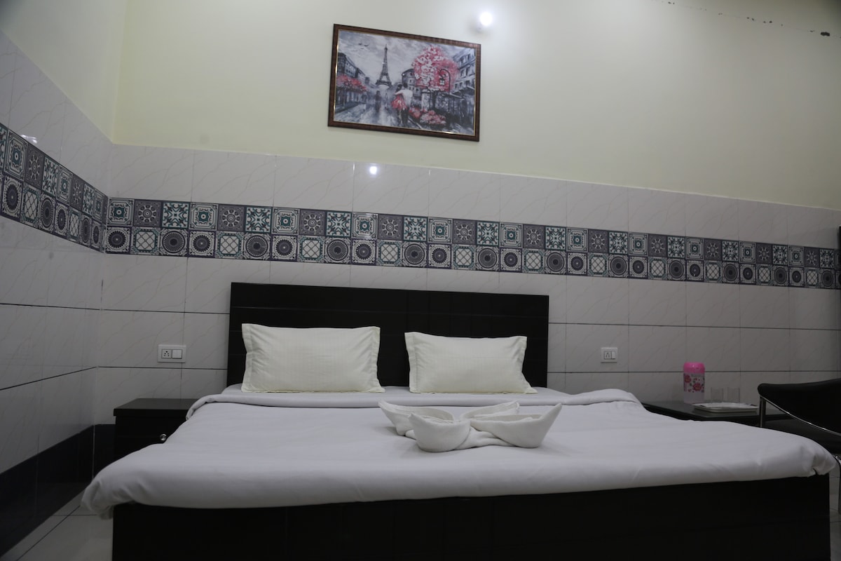 A spacious double bed is presented with crisp white linens, accompanied by a decorative wall painting above. The walls feature a patterned tile border, enhancing the room's aesthetic. A bedside table and a few personal items are visible, providing a sense of home within the inviting space.