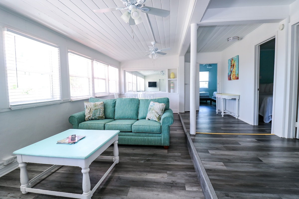 A light-filled living area is showcased, featuring a teal couch with decorative pillows. A soft blue coffee table is positioned in front. Beyond, a doorway leads to a bedroom, while large windows allow natural light to illuminate the space.