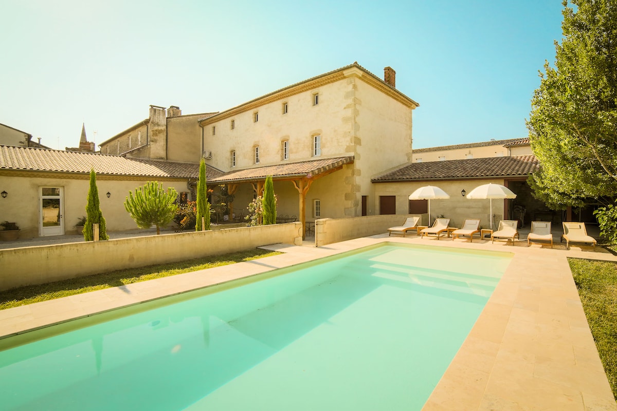 A sunny view of the spacious pool area, featuring a turquoise pool surrounded by a smooth stone deck. Sun loungers are positioned along the edge, under large umbrellas. The charming two-story house provides a serene backdrop, enhanced by surrounding greenery.