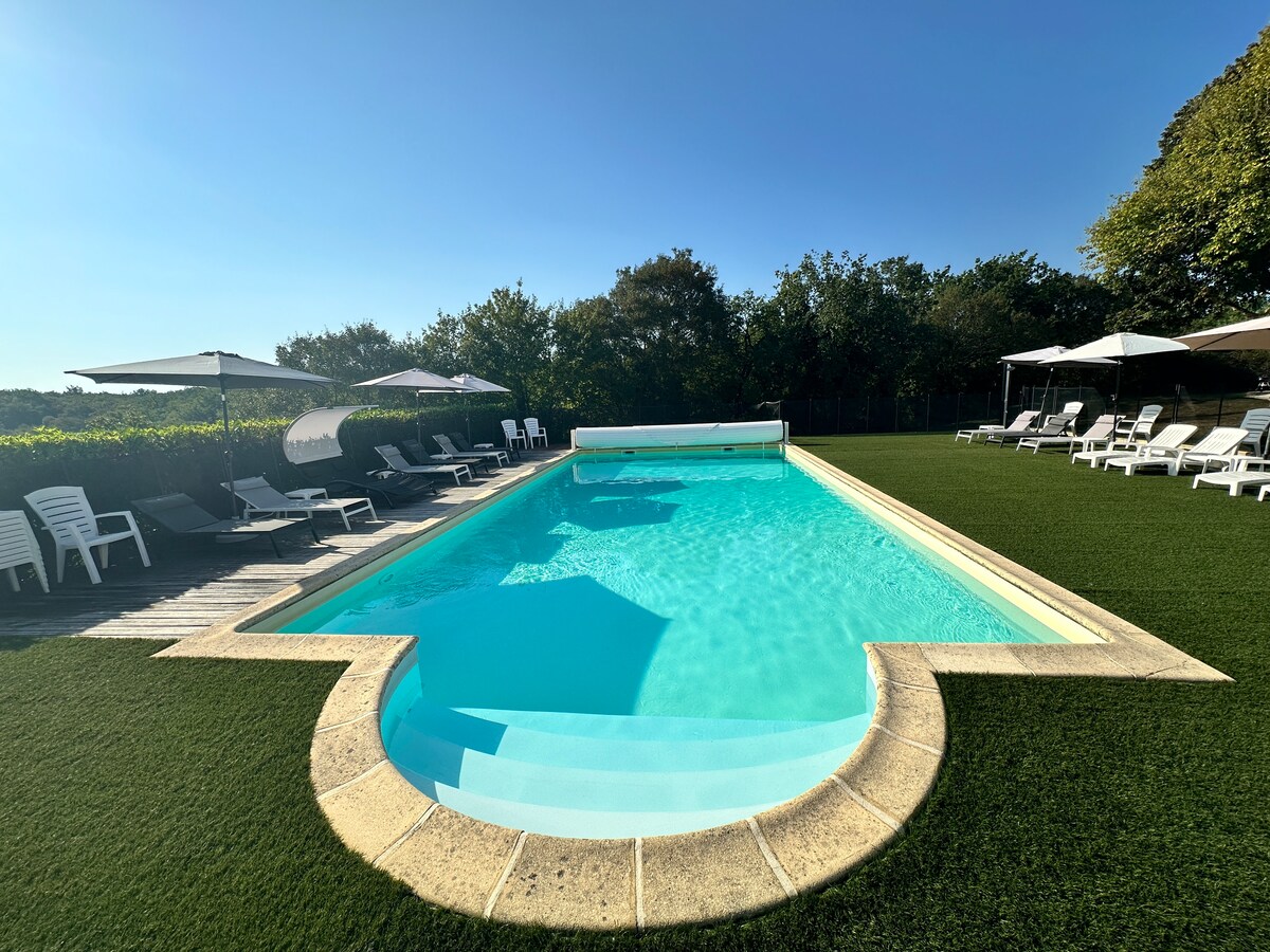 A glistening swimming pool is surrounded by lush green grass and equipped with several sun loungers. Shade is provided by large umbrellas, creating a serene space for relaxation. The backdrop features natural greenery, enhancing the tranquil atmosphere.
