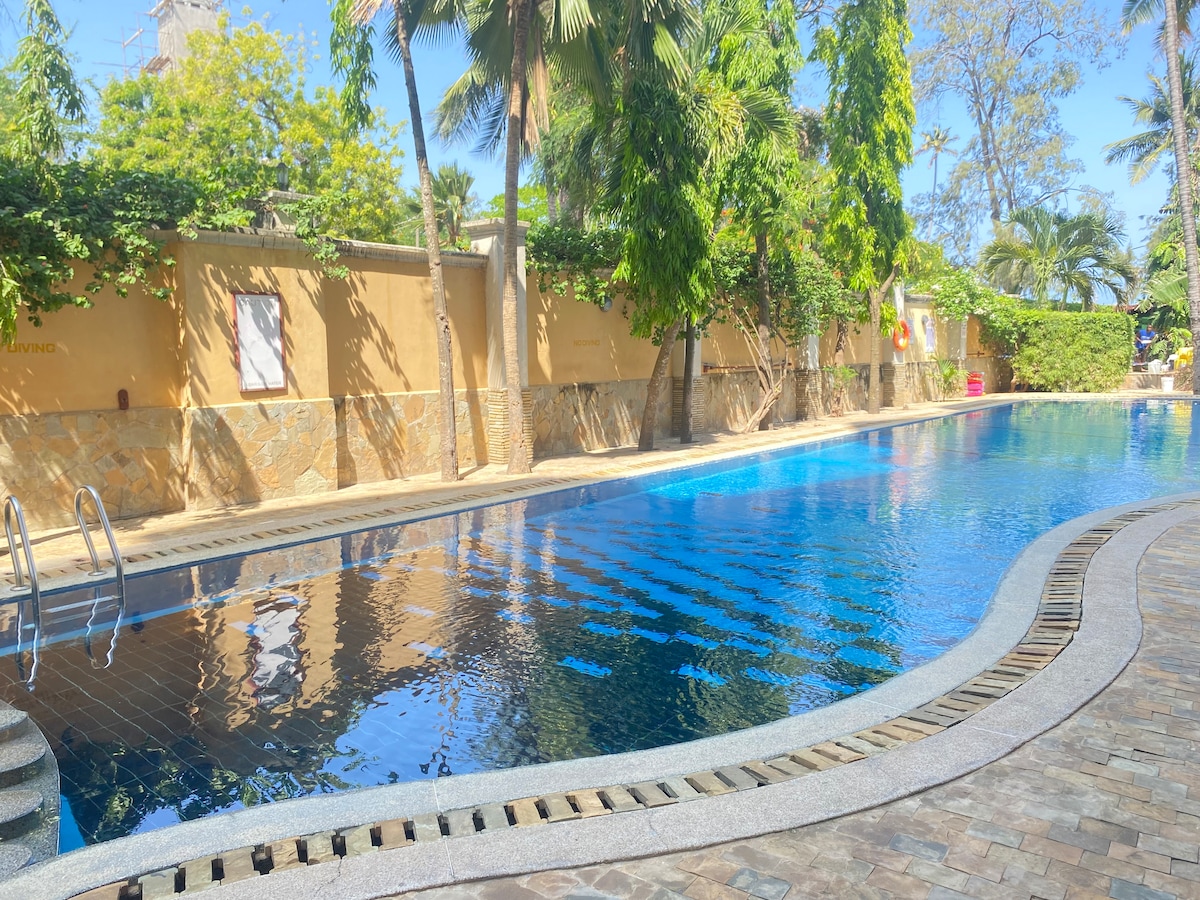 A large swimming pool is surrounded by lush greenery and tall palm trees. The pool features a gently curved edge with stone tiling leading to a set of stairs for easy entry. Sunlight reflects off the water, creating a calming atmosphere.