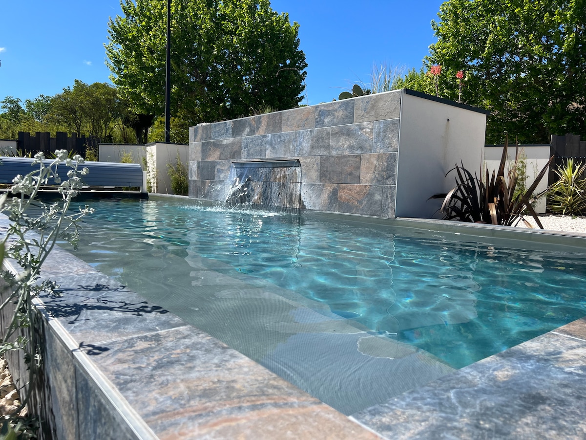 A modern pool is showcased with a tranquil water feature cascading over a stone wall. The pool’s clear water reflects sunlight, surrounded by lush greenery and tasteful landscaping. A serene ambiance is created by the gentle flow of water and the vibrant natural surroundings.