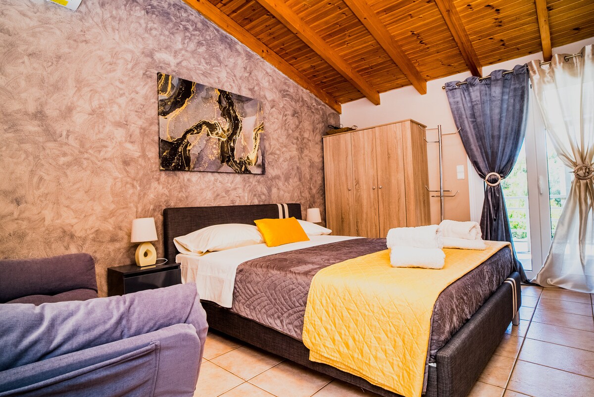 A spacious bedroom features a large bed dressed in neutral linens with an accent pillow. A soft throw blankets the foot of the bed. Natural light floods the room through glass doors, complemented by sheer curtains. A wooden wardrobe and a cozy armchair are also visible.