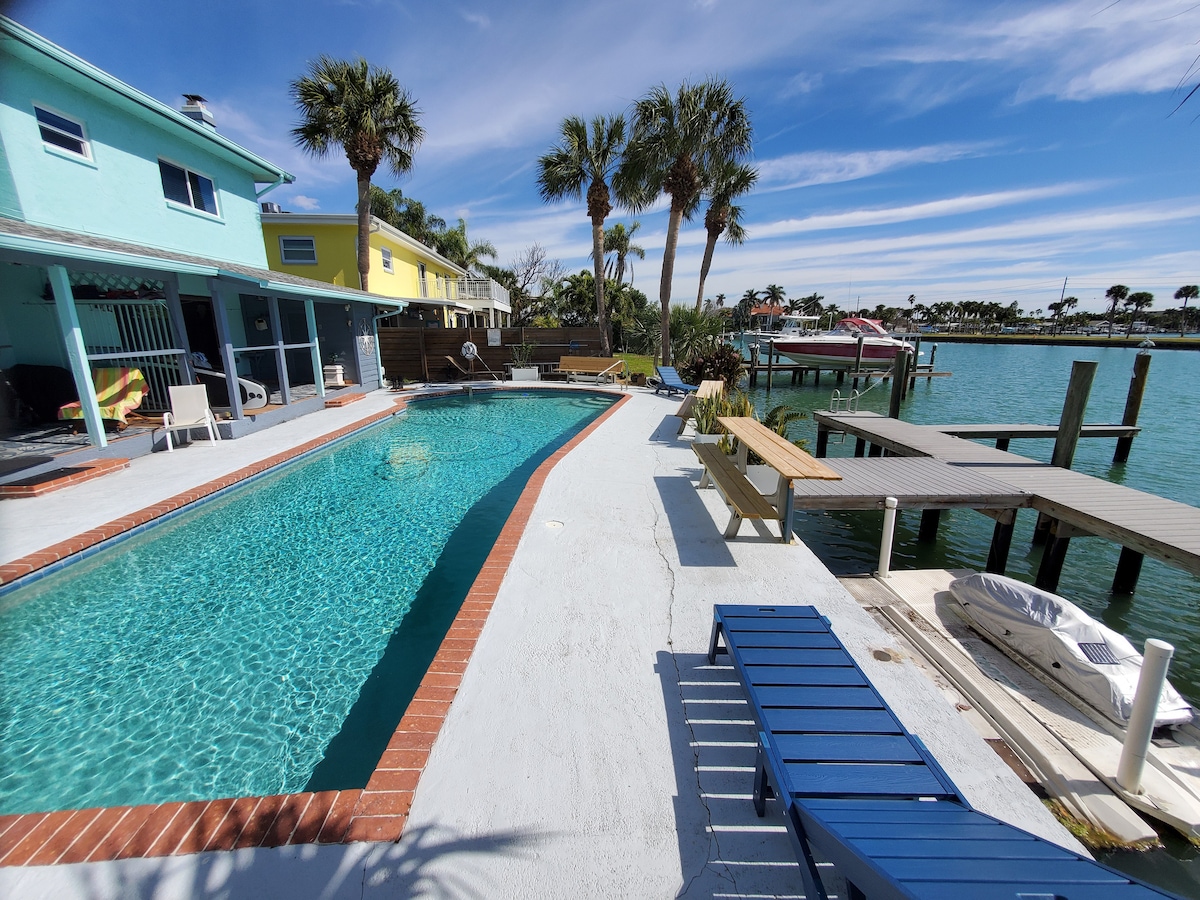 A clear pool is framed by a spacious deck, with lounge chairs positioned nearby. Palm trees line the water's edge, enhancing the coastal view. A wooden dock extends into the canal, featuring a boat. Colorful townhouses are visible in the background against a sunny sky.