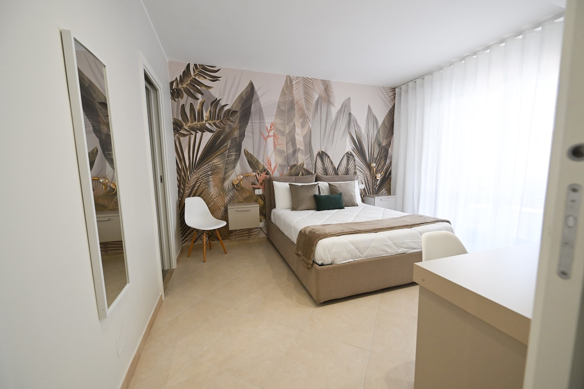 A modern bedroom features a spacious bed with neutral bedding, complemented by decorative pillows. A large wall mural depicts tropical leaves, adding a serene touch. Light filters softly through sheer curtains, illuminating the room. A minimalist chair and mirrored wardrobe complete the inviting space.