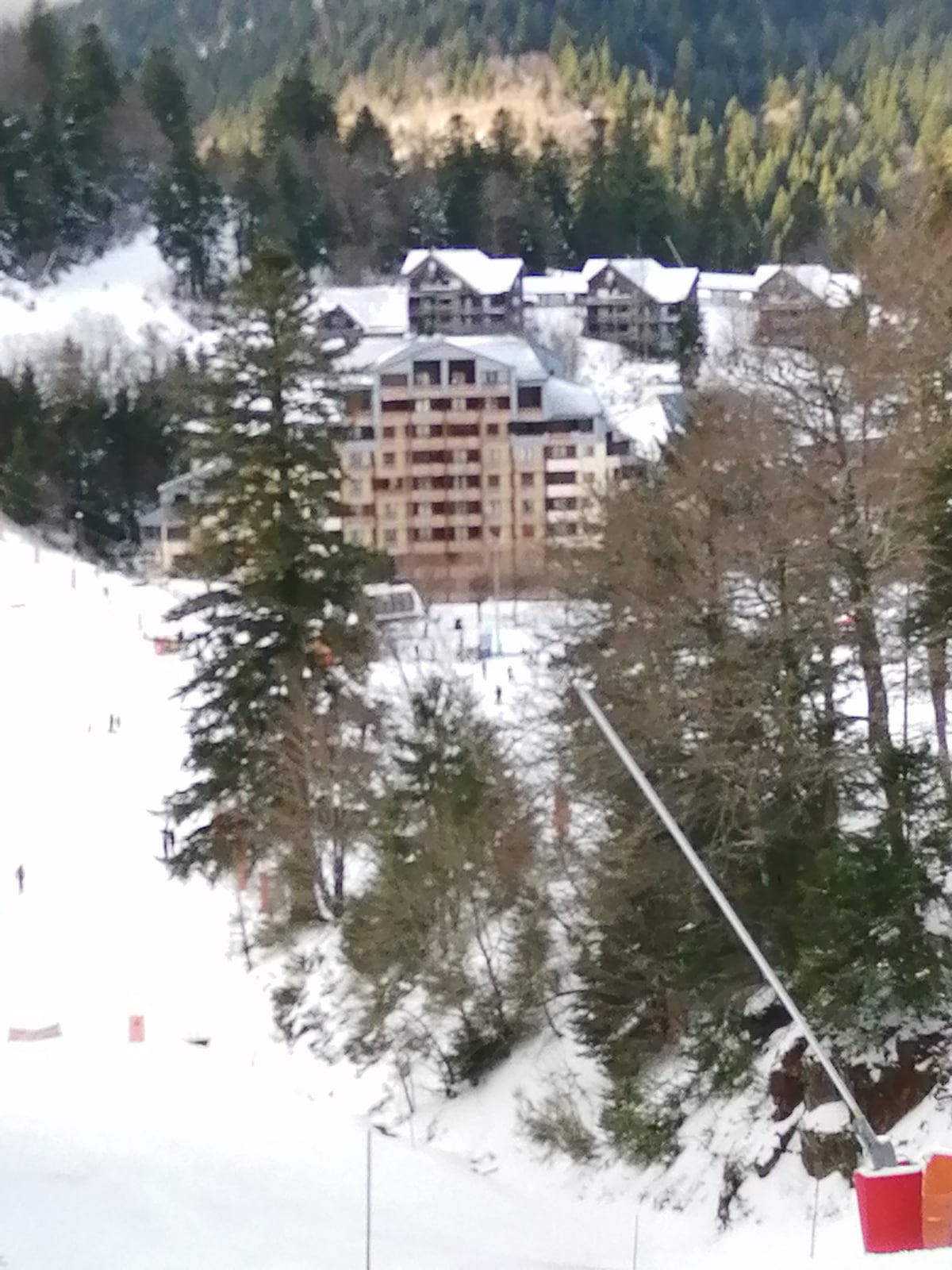 A multi-story building is seen nestled among coniferous trees, surrounded by snow-covered ground. The ski slopes extend below, indicating proximity to winter activities. The landscape features a mix of natural greenery and the structure's modern architecture, situated against a backdrop of distant mountains.