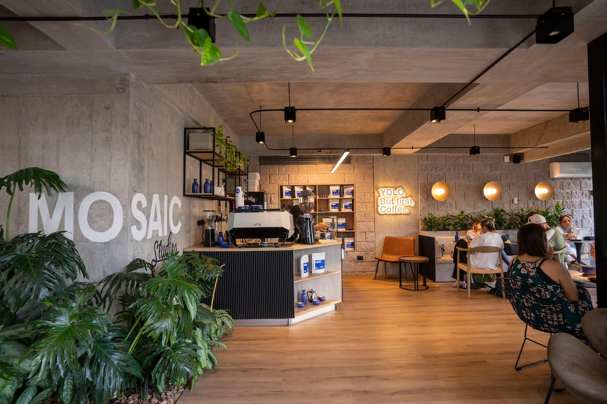 A modern coffee shop with a sleek counter displays various coffee-making equipment. Natural light illuminates the space, highlighting wood floors and vibrant greenery. Patrons are seated at different tables, enjoying their drinks in a relaxed atmosphere accented by contemporary lighting fixtures.