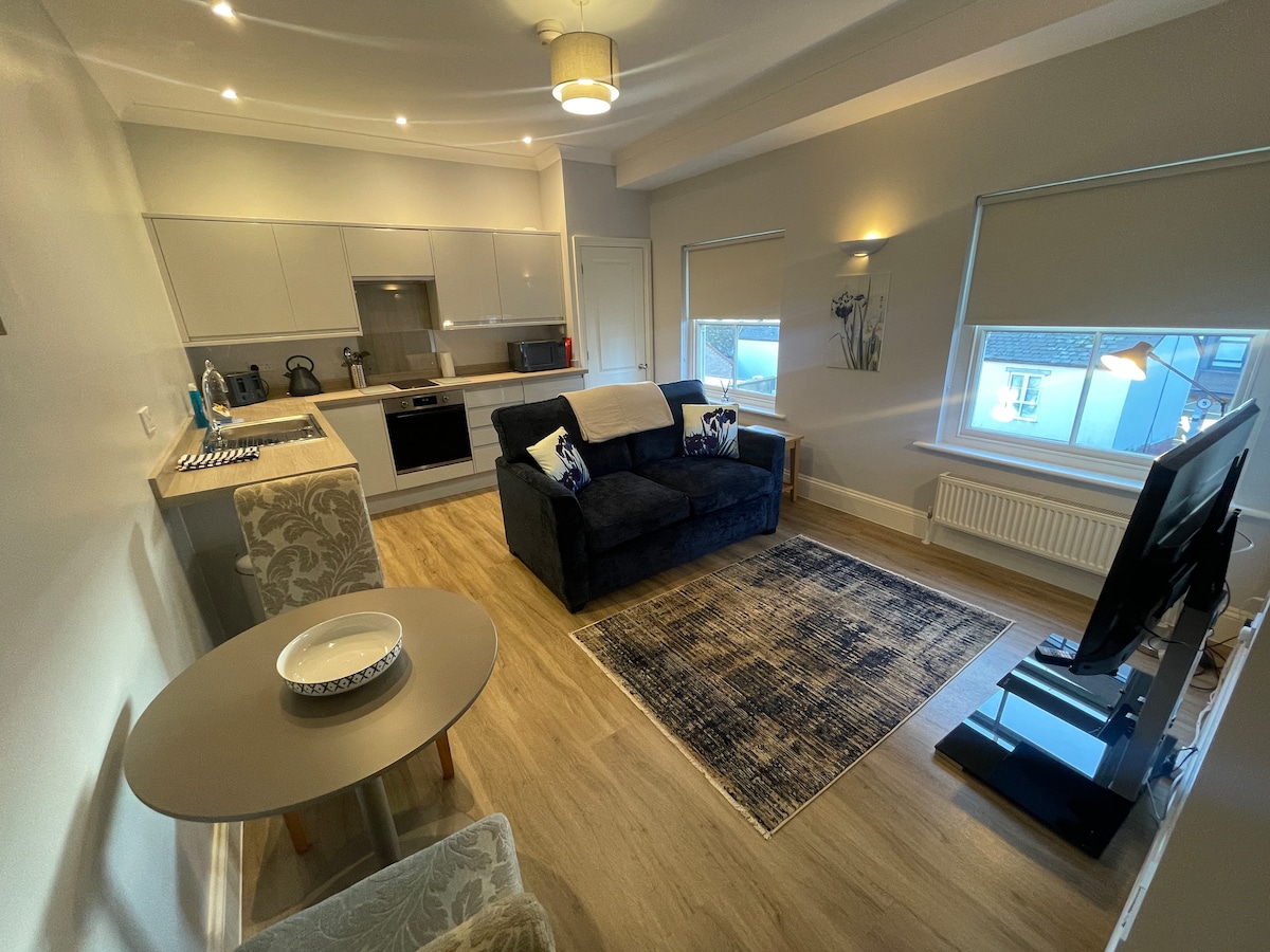 An open-plan living space is showcased, featuring a streamlined kitchen with modern appliances, including a microwave and oven. A neutral sofa sits opposite a wall-mounted TV. A circular dining table is present, with a decorative plate placed atop, while light enters through large windows.