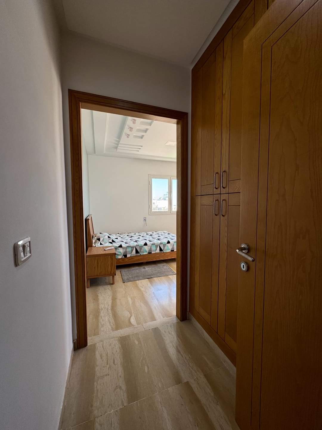 A hallway leads to a well-lit bedroom, featuring wooden cabinetry along one wall. The floor is finished with light-colored tiles, and a cozy bed is visible in the background, framed by a window allowing natural light to enter the space.