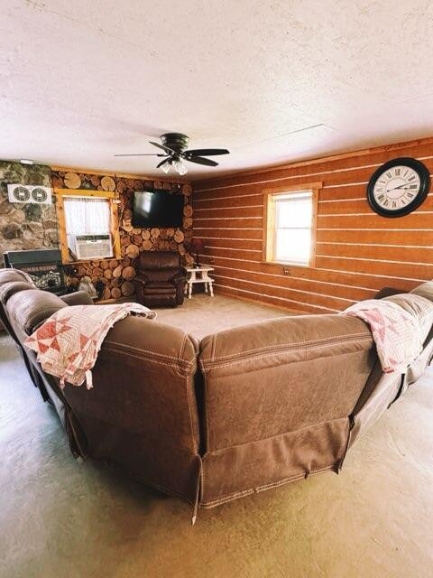 A spacious living area is furnished with a large sectional couch arranged in a circular formation. A ceiling fan is positioned above, and a wall clock hangs prominently. Natural wood accents are present on the walls, complementing the cozy atmosphere.
