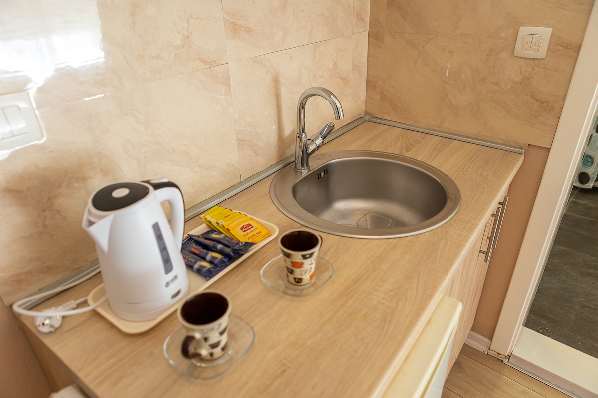 A compact kitchenette is shown, featuring a stainless steel sink and a kettle placed on a wooden countertop. Two cups are positioned on glass saucers beside a small selection of tea bags, all set against a backdrop of light-colored tiles.