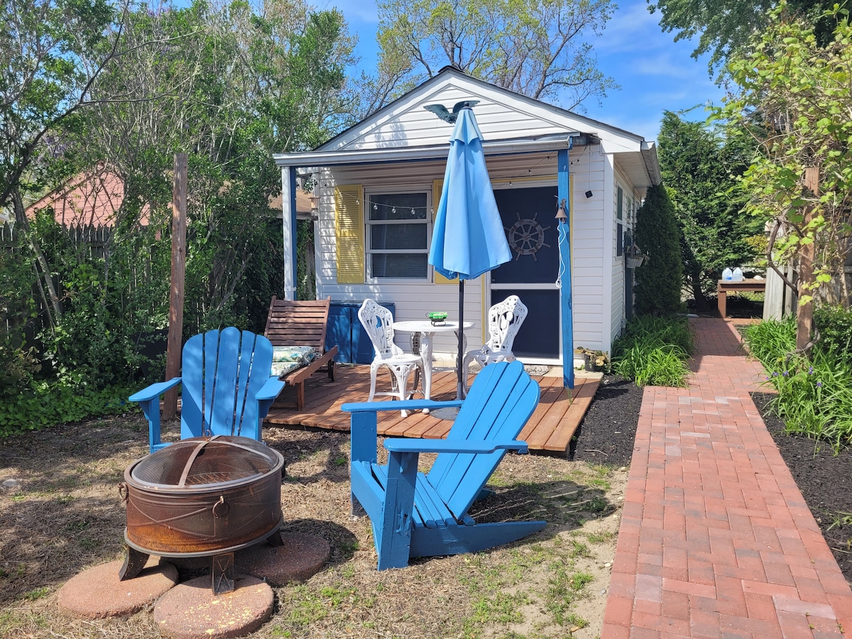 An outdoor seating area is presented with blue adirondack chairs arranged around a fire pit. A small table with matching chairs is positioned on a brick path leading towards the entrance of the dwelling, which is accented by greenery and a blue umbrella.