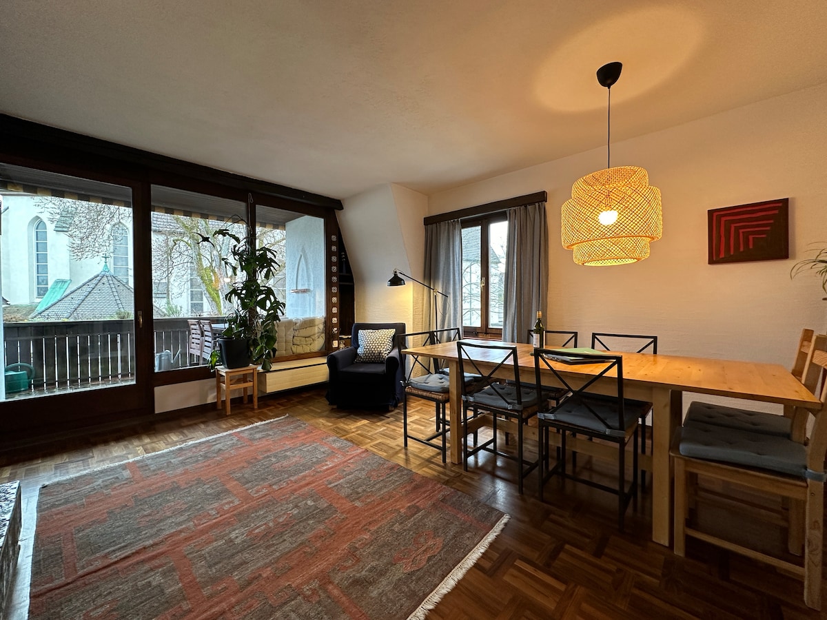 A spacious living and dining area is highlighted by large windows offering ample natural light. A wooden dining table with several chairs is arranged beside a cozy seating area featuring an armchair. Greenery from indoor plants adds a touch of brightness to the room's design.