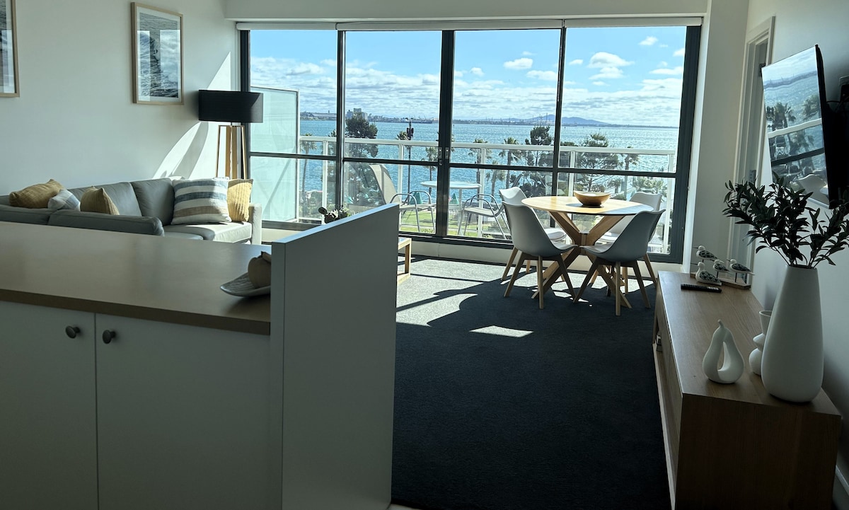 The living and dining area features large windows offering unobstructed waterfront views. A modern dining table with four chairs is positioned near a console table, and a cozy sofa is arranged for relaxation. Light filters in, creating an inviting atmosphere in the open space.