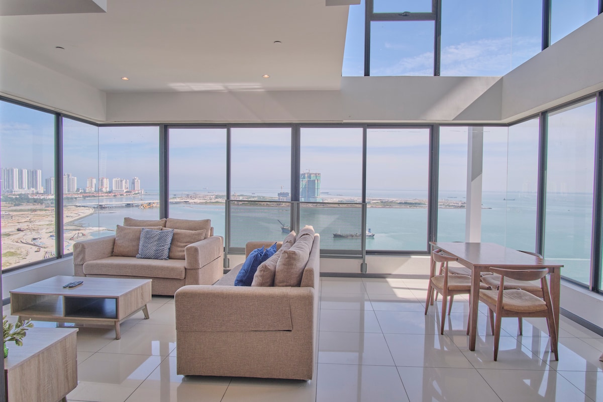A modern living area features large floor-to-ceiling windows that provide expansive views of the ocean and city. Comfortable sofas are arranged beside a sleek coffee table, while a dining area with a wooden table and chairs is positioned near the window, inviting natural light.