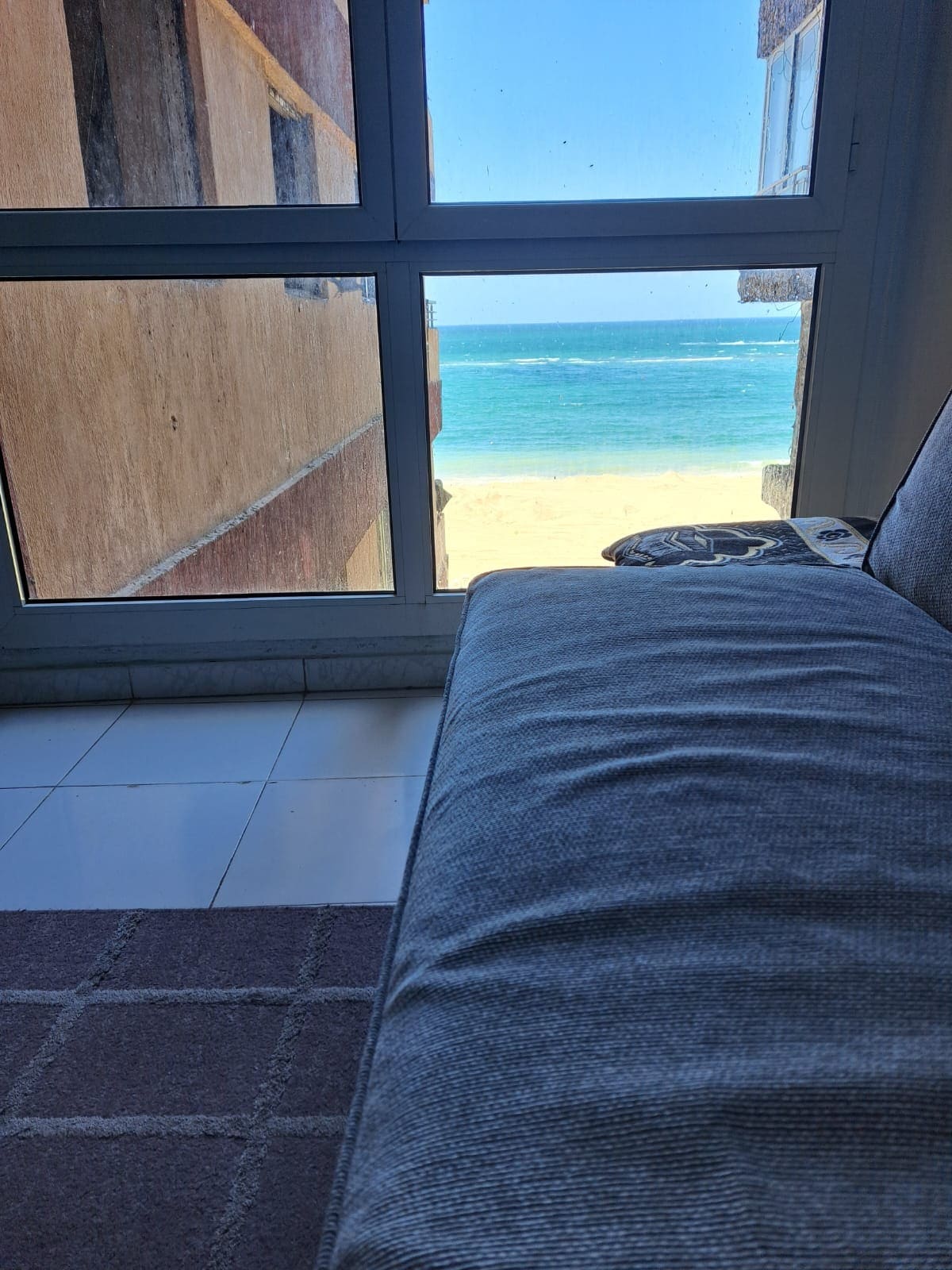 A cozy seating area features a large window offering a clear view of the beach and ocean. Soft natural light enters the space, illuminating the neutral tones of the furniture and the tiled floor, enhancing the coastal scenery just outside.