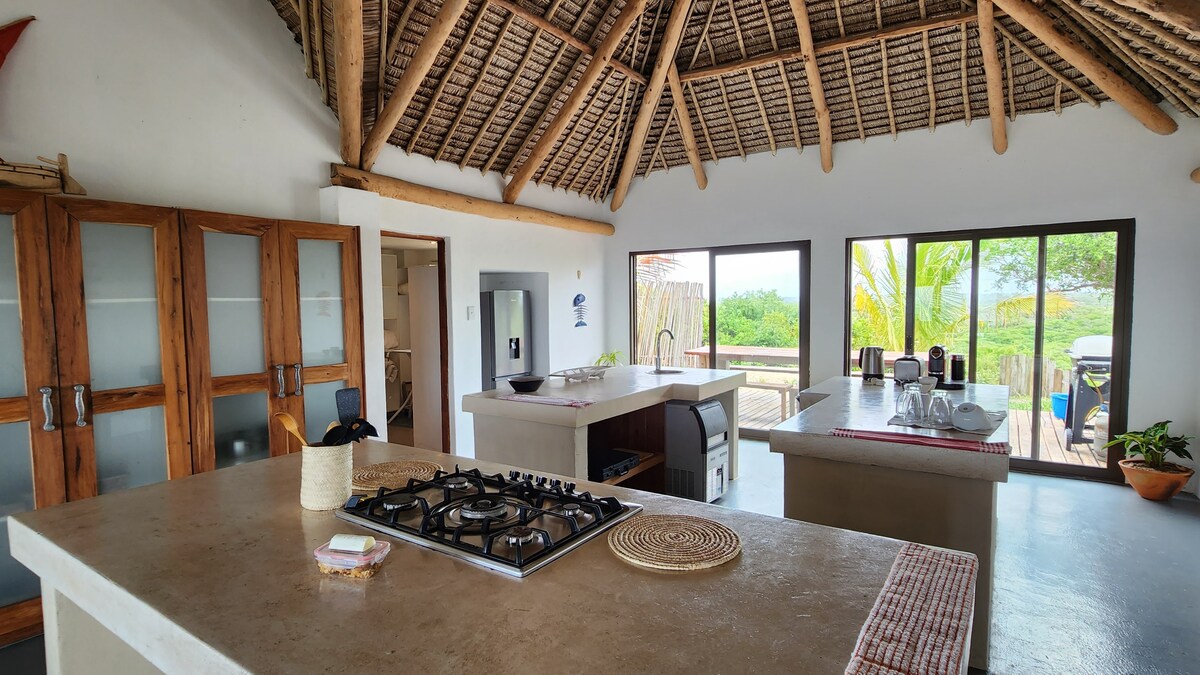 A spacious kitchen features an open layout with high thatched ceilings. Modern appliances include a gas stove and a refrigerator, complemented by ample counter space. Large windows allow natural light and provide views of the surrounding greenery.