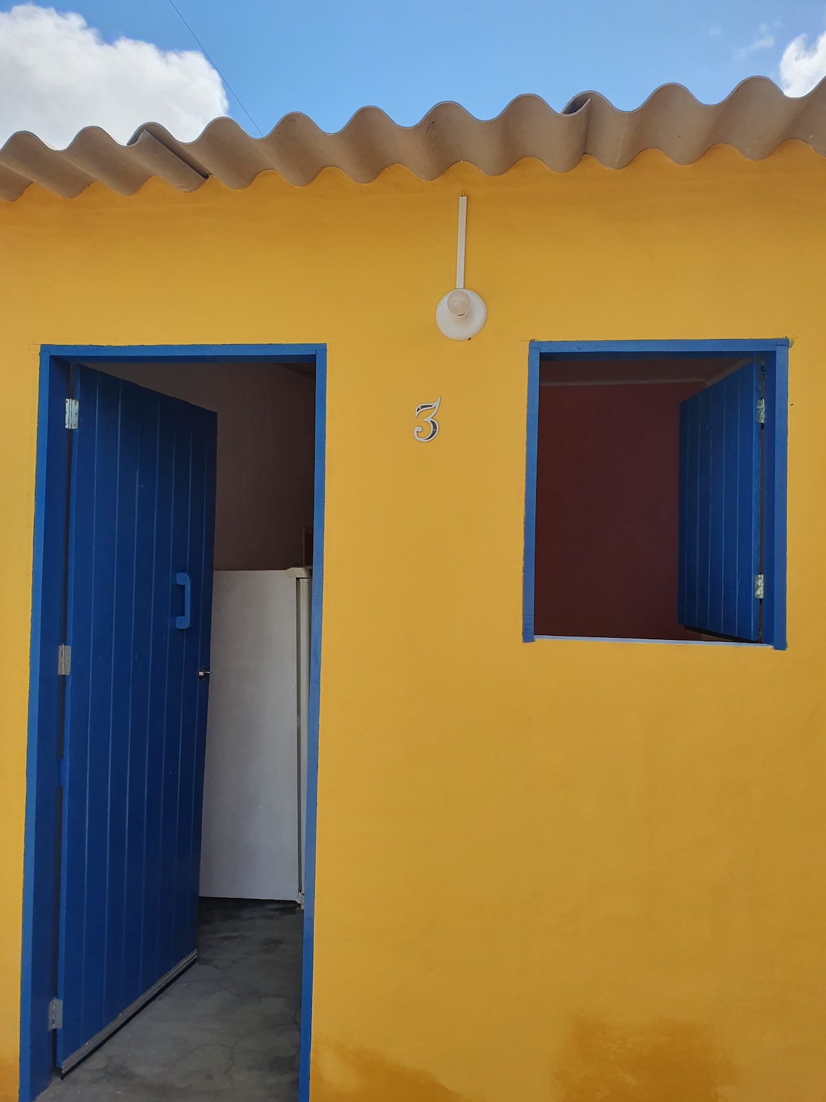 A vibrant yellow exterior is seen, featuring a blue door and a window. The number three is displayed above the door, and a small appliance is partially visible through the window, indicating the entrance to the space.
