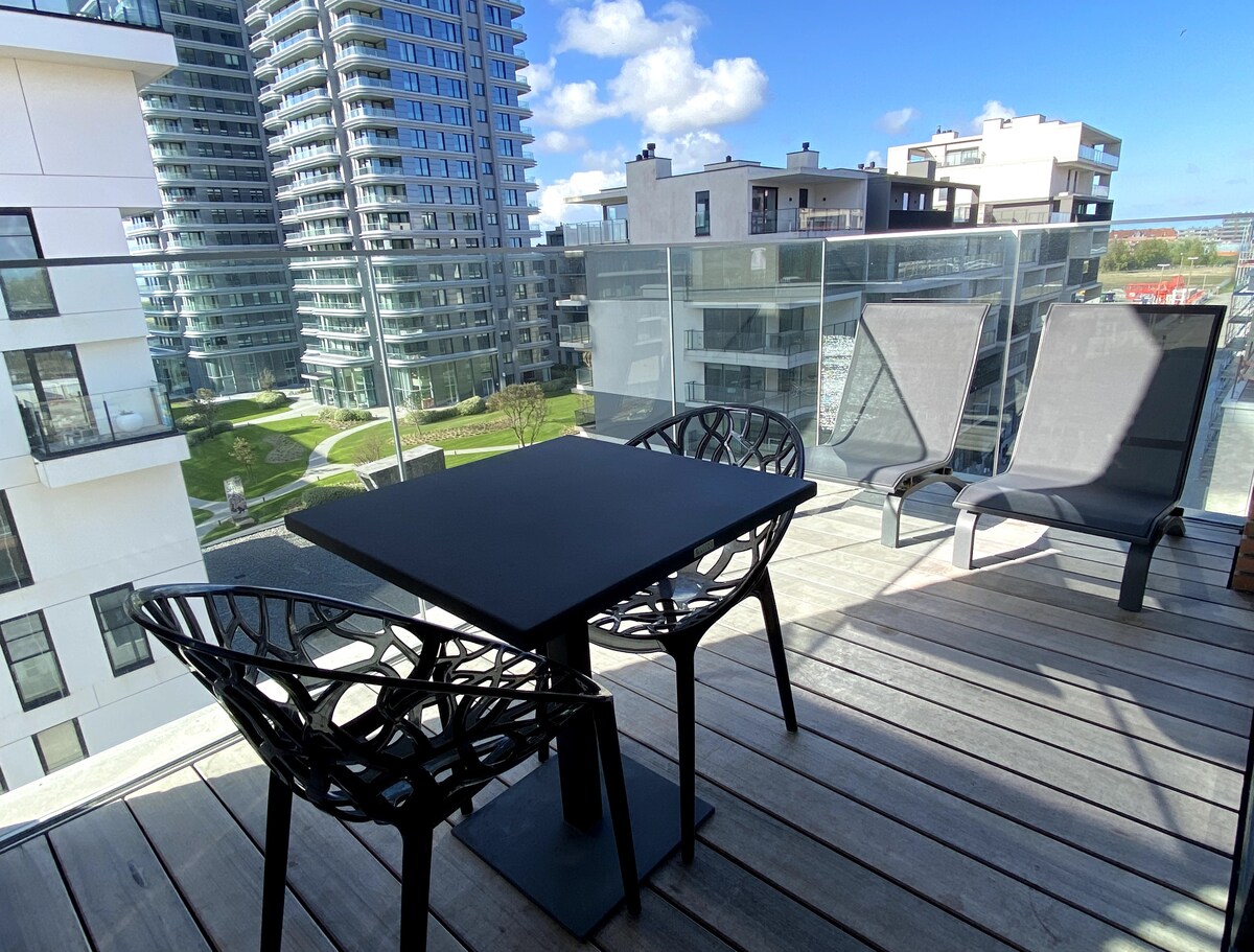 An outdoor terrace features a black table and two artistic chairs, providing a modern and comfortable space. A view of landscaped grounds and contemporary buildings can be seen in the background, framed by glass railings that enhance the open feel.