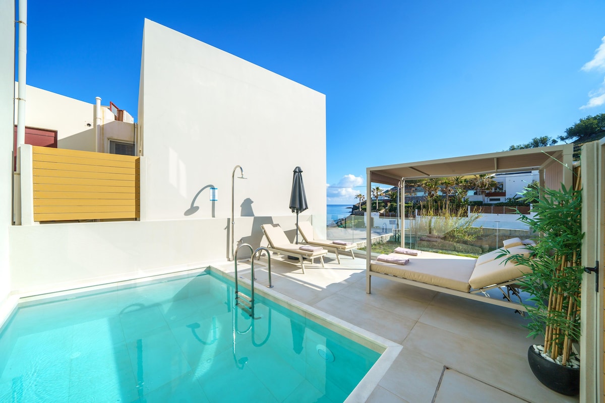 An outdoor space features a private swimming pool surrounded by light-colored tile. Two lounge chairs are positioned beside the pool, accompanied by a shaded cabana. The clear blue sky and greenery in the background enhance the open, welcoming atmosphere.