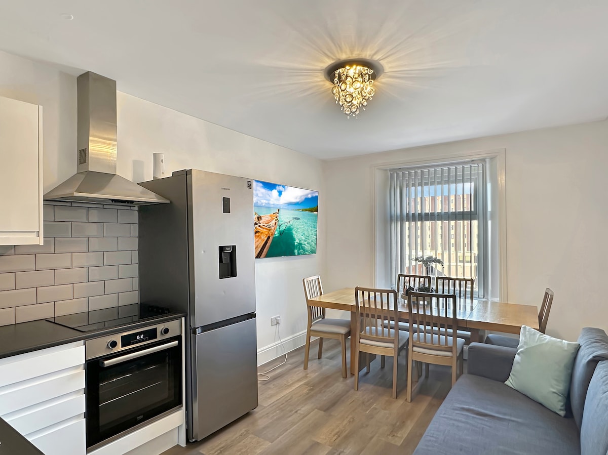 A modern kitchen and dining area is displayed, featuring sleek cabinetry and stainless steel appliances. A large refrigerator is paired with an oven and a stovetop. A dining table with four wooden chairs is positioned alongside a comfortable sofa, while a television mounted on the wall showcases a vibrant beach scene.