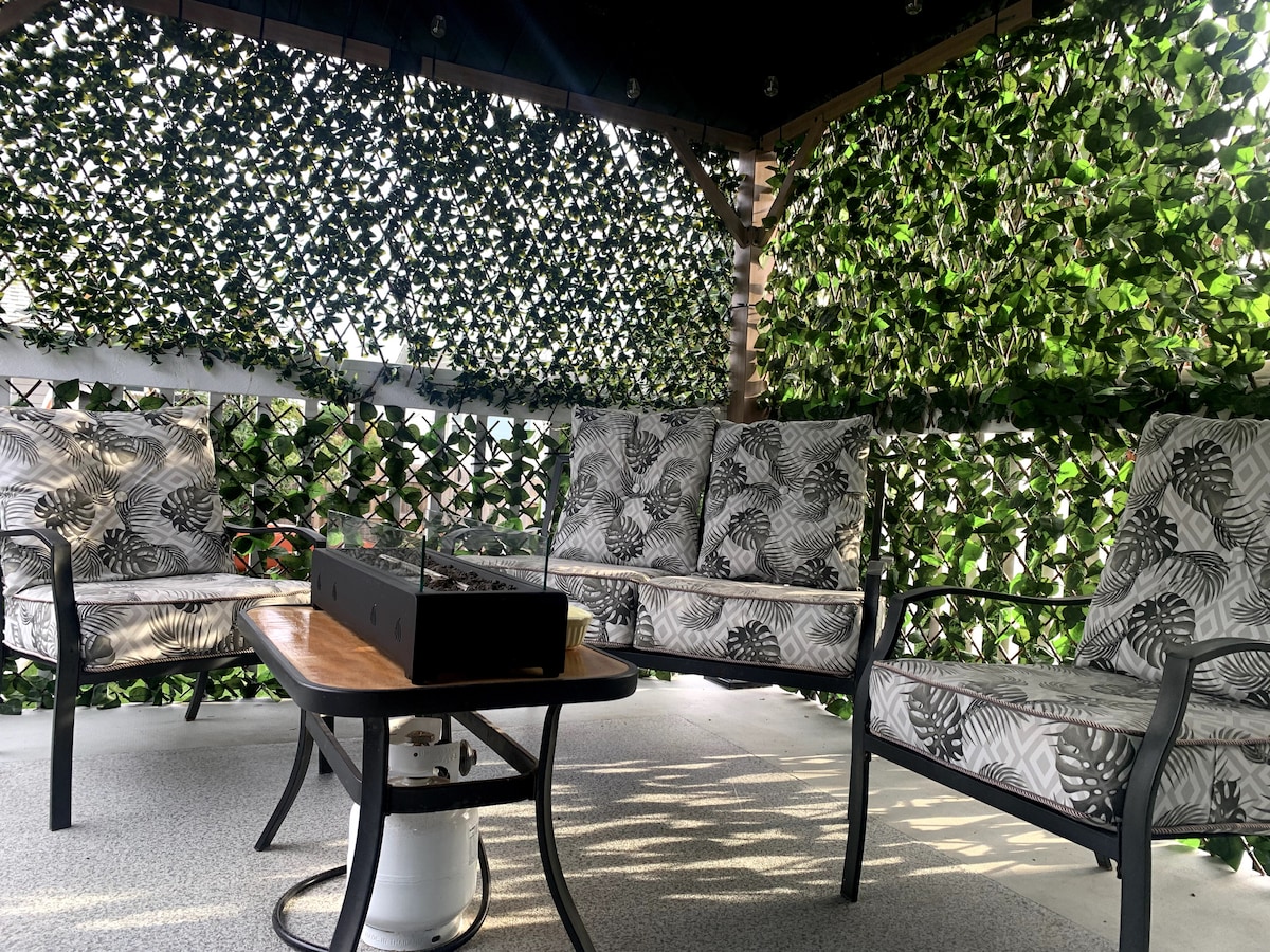 A secluded outdoor seating area is surrounded by lush green vines, creating a private and serene environment. The space features four cushioned chairs adorned with leaf-patterned fabric arranged around a wooden table, ideal for relaxation or gatherings.