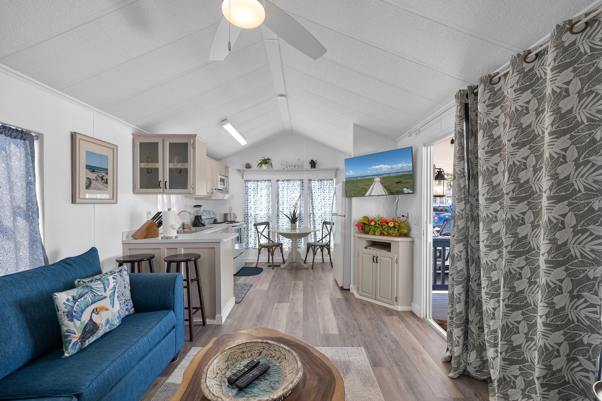 An open living area features a blue sofa with decorative pillows, accompanied by a wooden coffee table. A dining area with a round table and four chairs is visible, alongside large windows draped in light fabric. A kitchen with modern cabinetry is illuminated by natural light.