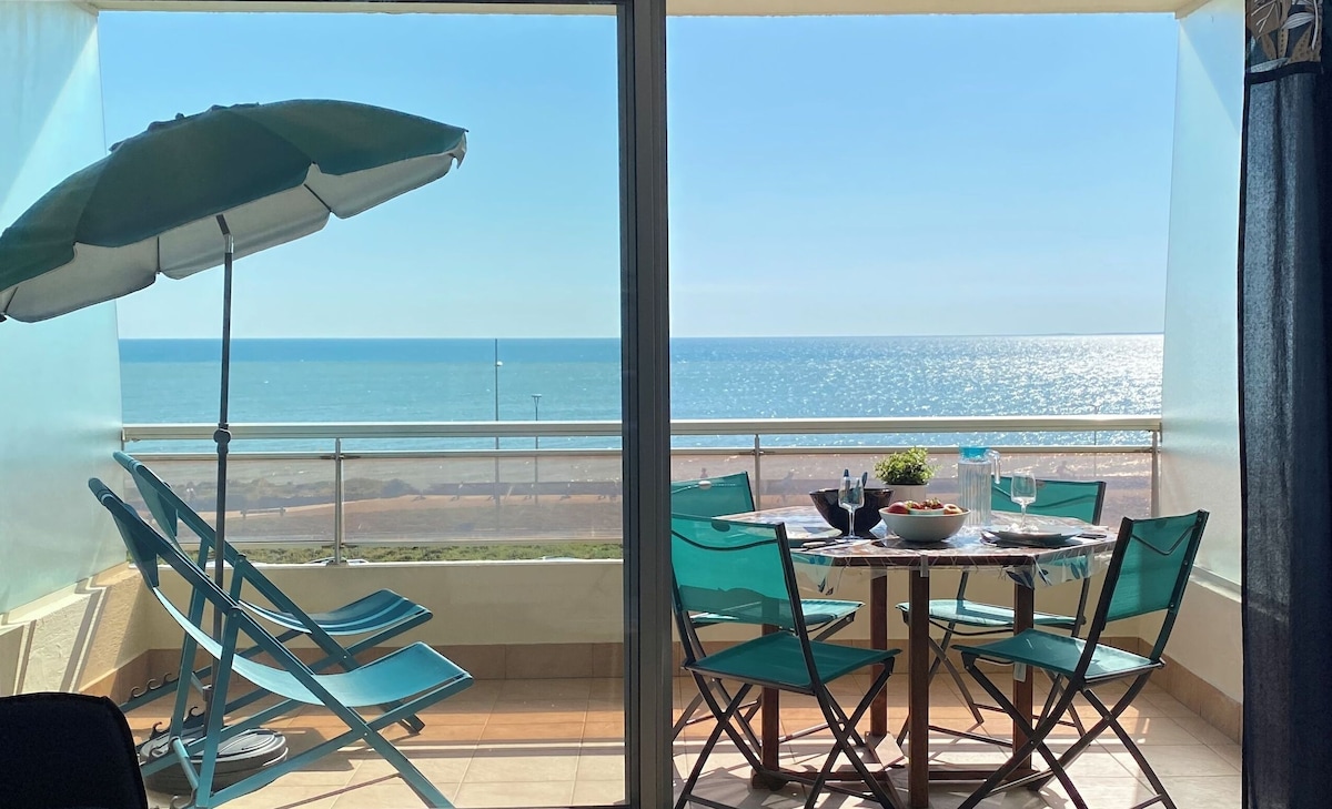 A spacious balcony features a round dining table surrounded by six teal chairs. A beach umbrella provides shade while two sun loungers are positioned for relaxation. The ocean glistens in the background under a clear sky, creating a serene coastal view.