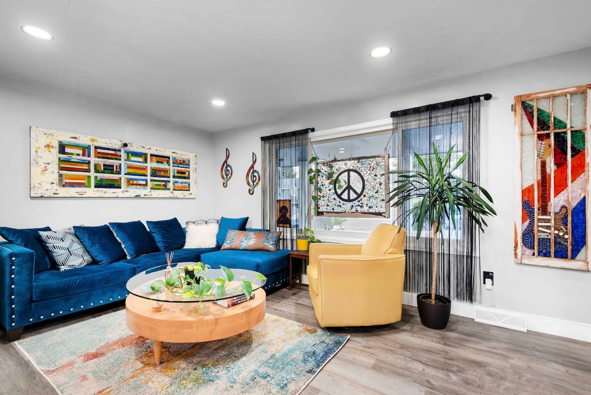 An inviting living room is presented, featuring a deep blue sectional sofa adorned with cushions, a round wooden coffee table, and a large glass window. Colorful artwork is displayed on the walls, complemented by a peaceful indoor plant and a yellow accent chair.