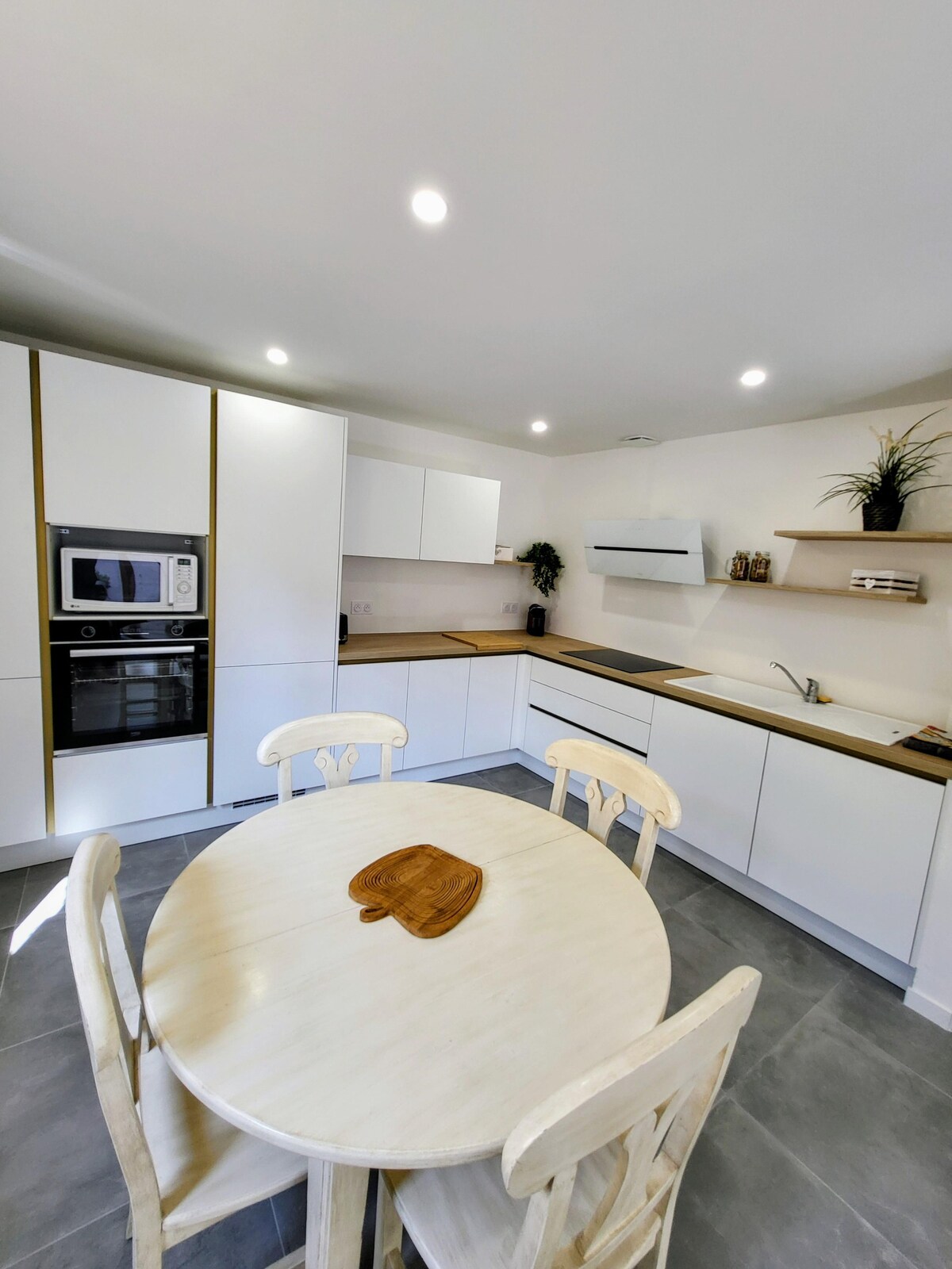 The kitchen showcases modern cabinetry with a mix of white and wood finishes. A round dining table is centered under recessed lighting, accompanied by two wooden chairs. An oven and microwave are built into the cabinetry, and countertops provide ample workspace.