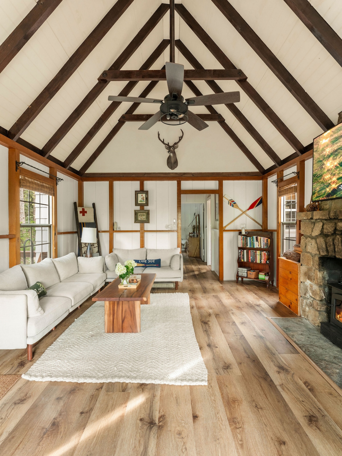 The living room showcases a light-colored sofa positioned across from a stone fireplace, with a wooden coffee table centered on a textured area rug. High ceilings are adorned with exposed beams, and natural light filters through multiple windows, illuminating the rustic decor.