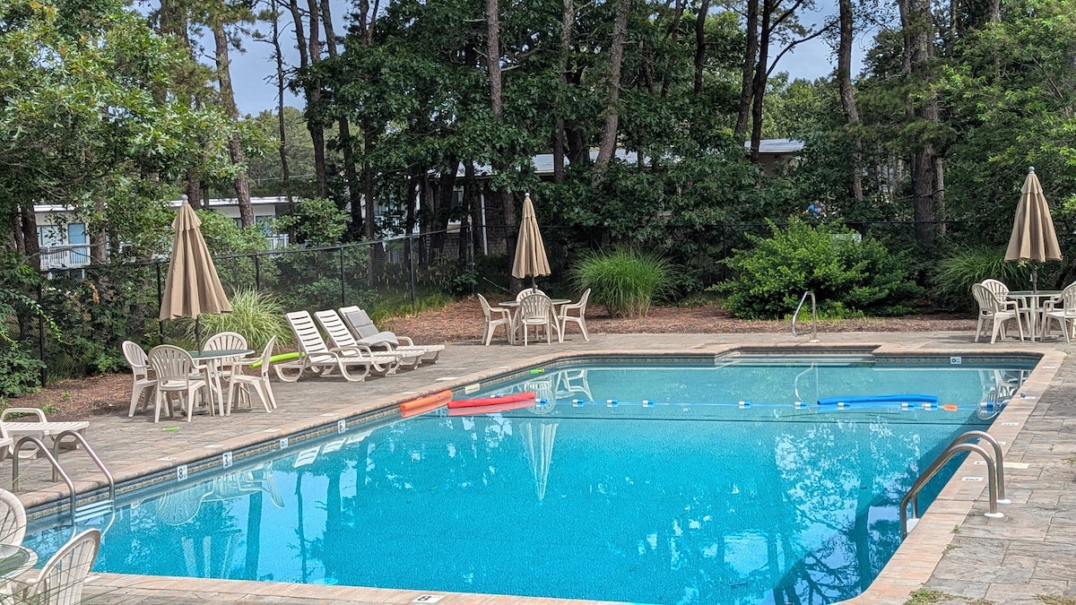 A community pool is surrounded by a stone patio, featuring lounge chairs, umbrellas, and a lush green landscape. The clear, blue water of the pool reflects the surrounding trees, creating a refreshing environment. Various inflatables float gently on the water's surface.