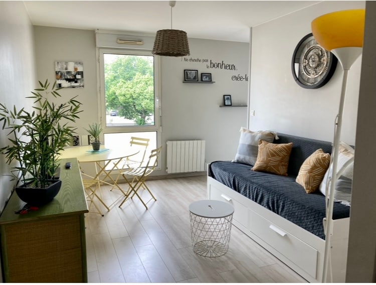 The living area is bright, featuring a comfortable sofa bed and a round coffee table. A small dining table with two folding chairs is present. Soft throw pillows add a touch of comfort, while greenery enhances the space. A large window provides natural light.