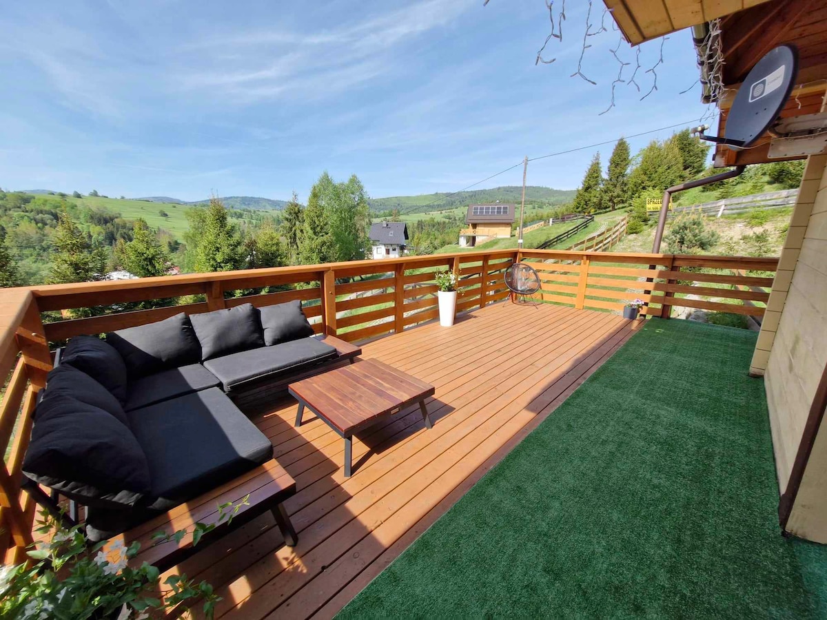 A spacious wooden deck features a comfortable black sofa and a low table, providing a relaxed outdoor seating area. Lush greenery and rolling hills are visible beyond the railing, enhancing the tranquil atmosphere. The deck is adorned with artificial grass and a potted plant.