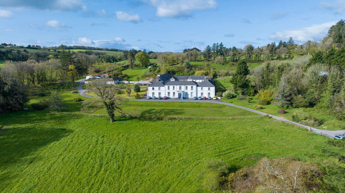 Aerial view of Myross Wood House showcases the historic structure surrounded by lush green fields and woodlands. The building, featuring a light exterior and dark roof, is set against a backdrop of rolling hills under a partly cloudy sky.