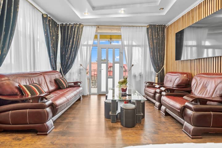 This inviting living room boasts a rich, classic aesthetic with a comfortable leather sofa, elegant drapery, and ample natural light, creating a relaxing space for gatherings or quiet moments.