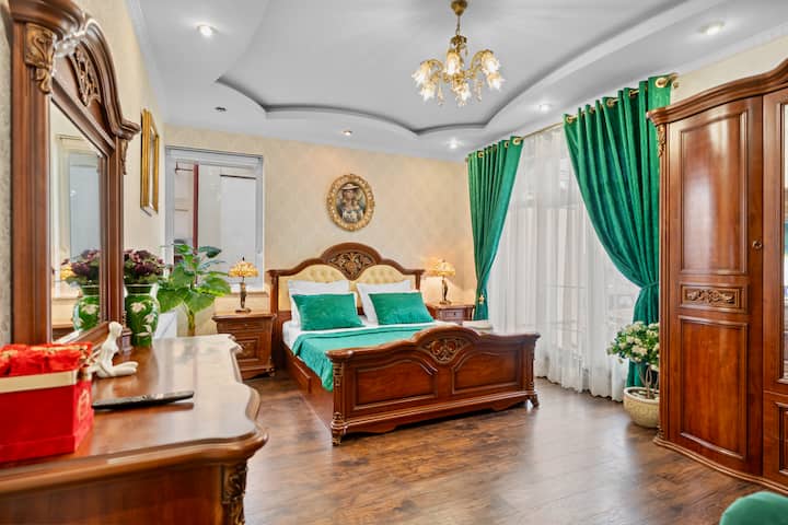This grand bedroom features a luxurious carved wood bed, elegant furnishings, rich textures like the emerald drapes, and a sparkling chandelier for a truly opulent experience.