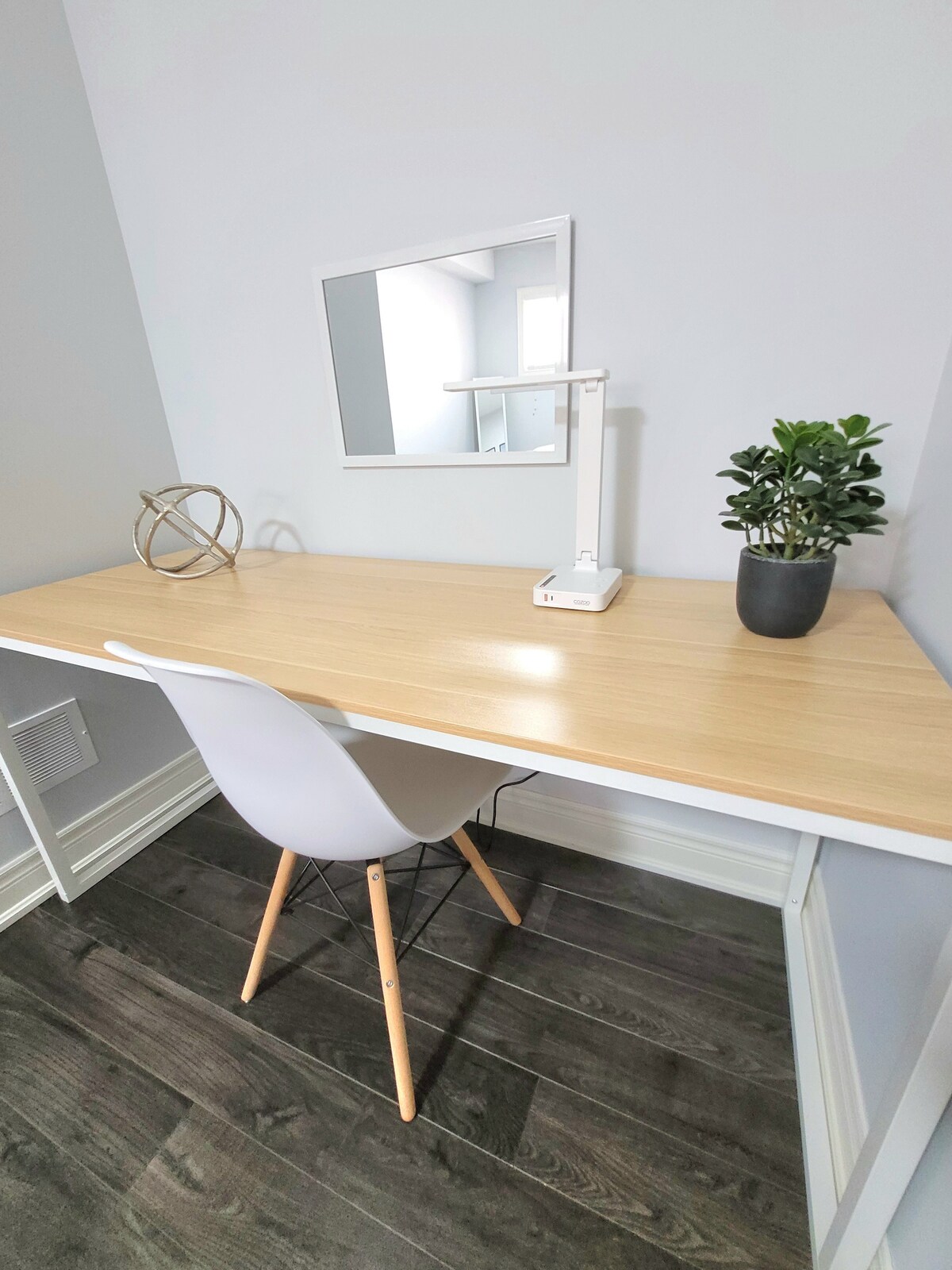 A minimalist workspace is featured with a light wood desk and a white chair. A small plant in a black pot and a geometric decorative piece are placed on the desk. A mirror is positioned above the desk, reflecting the natural light in the room.