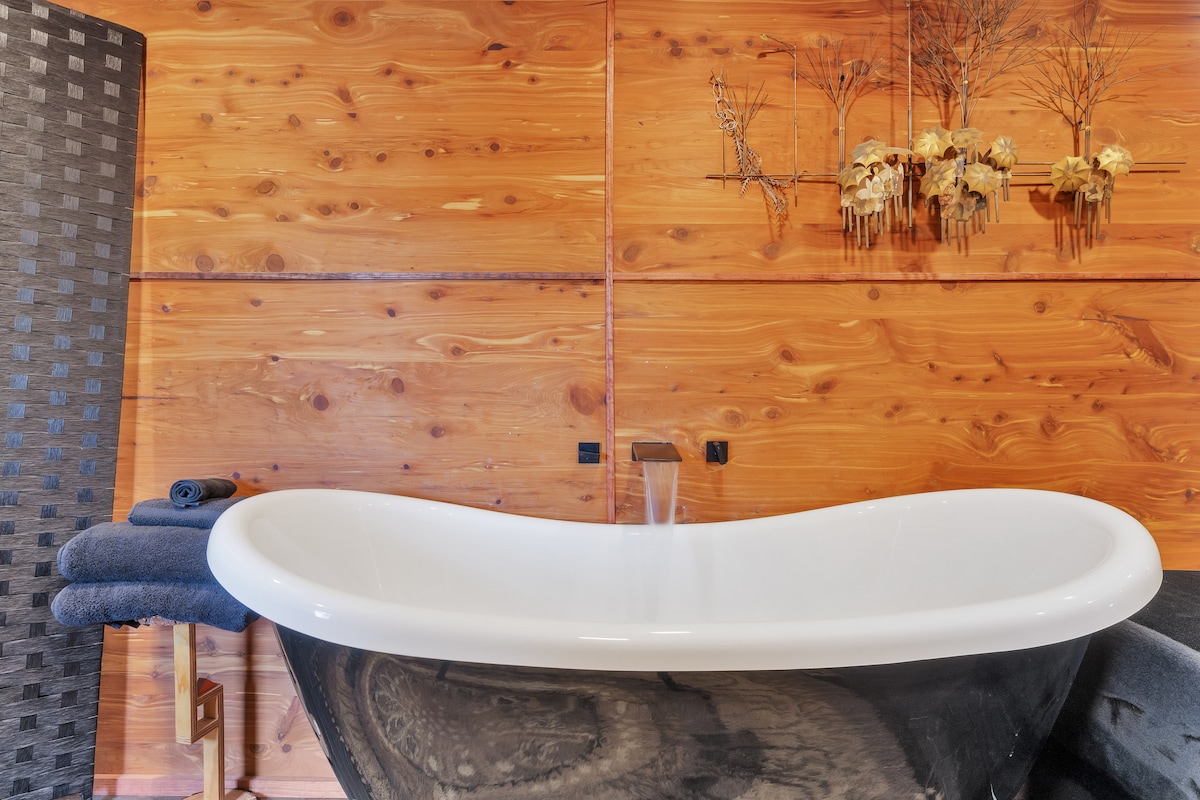 A sleek clawfoot bathtub is positioned against a backdrop of warm wooden paneling. Minimalist decor features a collection of skulls and dried flowers mounted on the wall. Soft gray towels are neatly arranged beside the tub, enhancing the tranquil atmosphere.